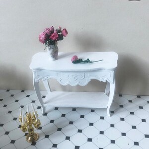 Peut inclure: Table console miniature blanche avec un motif sculpté sur l'étagère supérieure. La table a une seule étagère et repose sur un sol à motifs en losanges noirs et blancs. Un bouquet de fleurs roses dans un vase blanc et un chandelier doré avec des bougies blanches sont sur la table.