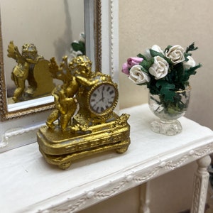 Op de afbeelding: Een miniatuur witte tafel met een goudomrande spiegel en een gouden klok met een engelenbeeldje. Een kleine vaas met bloemen staat op de tafel. De spiegel reflecteert de klok. De tafel heeft decoratieve randen.