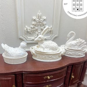 Könnte beinhalten: Miniatur-Keramikfiguren in Weiß: Huhn, Hase und Schwan mit Deckeln, auf einem dunklen Holzschrank. Der Schrank hat zwei Schubladen und eine kleine Tür mit goldfarbener Hardware. Der Hintergrund umfasst weiße Wanddekoration und ein rundes Logo mit der Aufschrift "Uptown Minis".