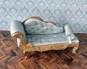 May include: A miniature blue chaise lounge with gold accents and a white pillow with blue trim. The chaise lounge is sitting on a wooden floor with a herringbone pattern. The background is a blue and white damask wallpaper.