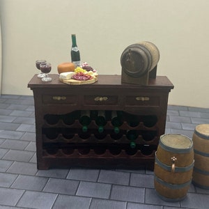 May include: A miniature wooden wine rack with a brown finish, featuring a wine bottle, two wine glasses, a cheese plate, and a wooden barrel. The rack is set on a gray tiled floor. The image includes the text "Uptown Minis".