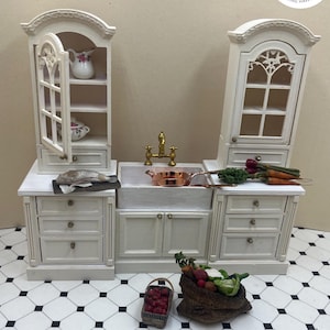 Puede incluir: Miniatura de cocina blanca con fregadero, armarios y una olla de cobre. El conjunto incluye una cesta de manzanas y una bolsa de verduras. El suelo está embaldosado con un patrón blanco y negro. El logotipo "Uptown Minis" está en la esquina superior derecha.