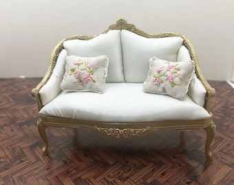 May include: A miniature dollhouse sofa with a gold-painted frame, white upholstery, and two small square throw pillows featuring pink floral embroidery. The logo Uptown minis is visible in the upper right corner of the image.