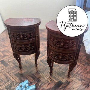 May include: Two miniature wooden nightstands with ornate carvings and two drawers each. The nightstands are on a wood floor with two blue miniature shoes.