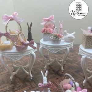 Puede incluir: Una escena de Pascua en miniatura con tres mesas blancas, cada una con una cesta de Pascua en miniatura diferente, un conejito y un arreglo floral. Las mesas están decoradas con encaje y tienen un acabado blanco desgastado. Las cestas están llenas de huevos en miniatura y flores. Los conejitos están hechos de madera y están pintados en un color madera natural. Las flores están hechas de tela y son rosas y blancas. La escena está ambientada en un suelo de madera con un acabado marrón claro. El texto "Uptown Minis" es visible en la esquina superior derecha de la imagen.