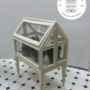 May include: A miniature white greenhouse with a glass roof and sides, containing small potted plants. The greenhouse features decorative scalloped edges and stands on four slender legs. The image includes the text "Uptown Minis" and "Crafting Happiness".