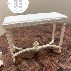 Puede incluir: Mesa consola en miniatura pintada de blanco con un diseño tallado en la parte superior y las patas. La mesa tiene un diseño simple y elegante y sería una gran adición a una casa de muñecas o una colección en miniatura.