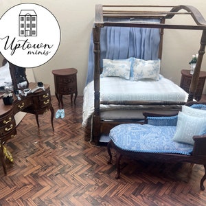 May include: A miniature dollhouse bedroom set with a canopy bed, a vanity with a mirror, two nightstands, and a chaise lounge. The bed is made with blue and white bedding, and the chaise lounge is covered in a blue and white paisley fabric. The furniture is made of wood and has a dark brown finish.