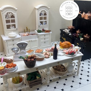 Peut inclure: Scène de cuisine miniature avec une table blanche chargée de nourriture, dont un poulet rôti, des légumes et du pain. Un poêle noir et des armoires blanches sont en arrière-plan. Une balance et une bouteille de vin sont sur la table. La scène est une cuisine de maison de poupée.