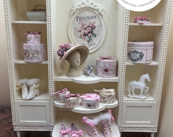 May include: A cream-colored miniature display cabinet featuring shelves filled with small decorative items including round patterned boxes, a mannequin head with a hat, a white horse figurine, and miniature shoes with pink bows.