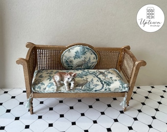 May include: A miniature gold-painted wooden sofa with a cane back and seat cushions. The cushions are covered in a floral fabric with a blue and white pattern. A small ceramic cat figurine is sitting on the sofa.