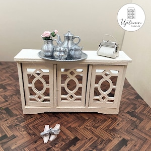 May include: A white miniature cabinet with three glass doors and a silver tea set with a floral arrangement on top. The cabinet is sitting on a brown wood floor. The text "Uptown Minis" is visible in the upper right corner of the image.