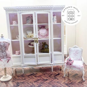 May include: White dollhouse-sized display cabinet with glass doors and shelves. The cabinet is decorated with pearl-like beads. The cabinet is filled with miniature items, including hats, boxes, and other decorative items. A miniature chair with a floral pattern and a miniature dress form with a pink lace dress are in front of the cabinet.