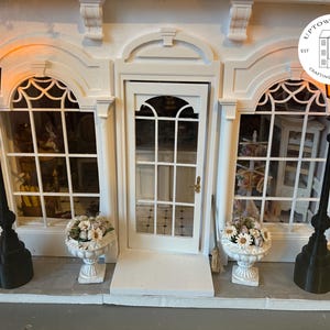 Puede incluir: Una fachada de edificio en miniatura blanca con una puerta central, dos ventanas y adornos decorativos. Dos farolas negras con luces naranjas brillantes flanquean las ventanas. Jarrones blancos con flores se encuentran debajo de las ventanas. Incluye el texto "Uptown Minis" y un logotipo.
