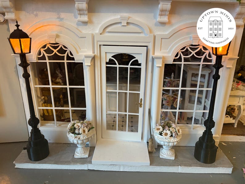 Puede incluir: Una fachada de edificio en miniatura blanca con una puerta central, dos ventanas y adornos decorativos. Dos farolas negras con luces naranjas brillantes flanquean las ventanas. Jarrones blancos con flores se encuentran debajo de las ventanas. Incluye el texto "Uptown Minis" y un logotipo.