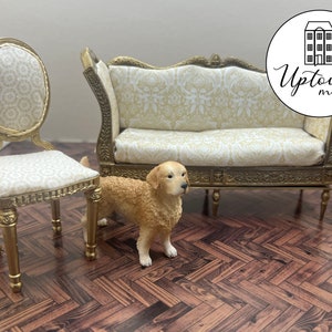 Könnte beinhalten: Ein Miniatur-Sofa in Gold mit weiß-goldener gemusterter Polsterung, ein passender Stuhl und eine Golden-Retriever-Figur. Die Möbel stehen auf einem braunen Holzboden mit Fischgrätenmuster.