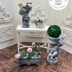 May include: Three miniature garden planters with cherub statues and pink flowers. The planters are on a white table with a white background. The text "Uptown Minis Est 2021 Crafting Happiness" is on a white circle.