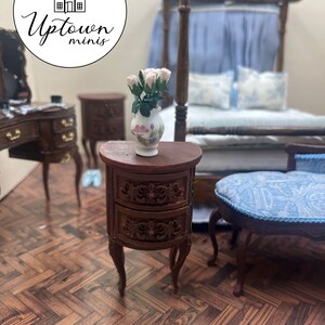 May include: A miniature wooden nightstand with two drawers and carved details. The nightstand has a curved front and sits on four legs. A vase with pink and white roses sits on top of the nightstand.