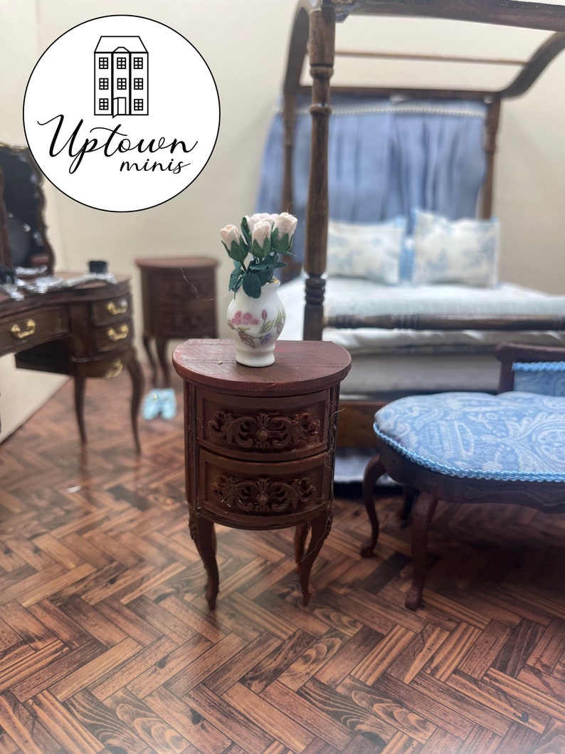 May include: A miniature wooden nightstand with two drawers and carved details. The nightstand has a curved front and sits on four legs. A vase with pink and white roses sits on top of the nightstand.