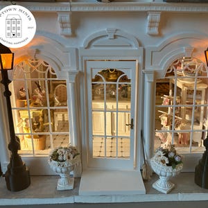 Puede incluir: Una fachada de tienda blanca en miniatura con una puerta central y dos grandes ventanas, flanqueada por farolas negras con luces naranjas. Jarrones blancos con flores en la base. La tienda tiene el texto "Uptown Minis" y "Crafting Happiness".