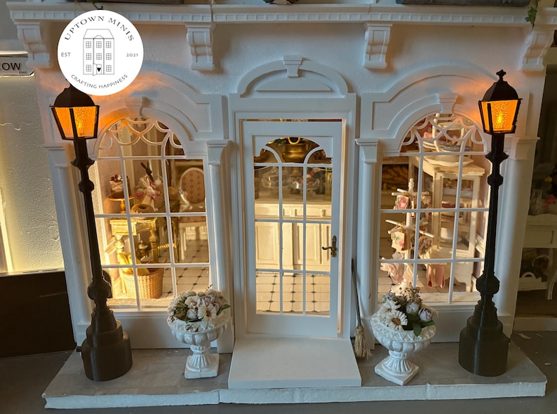 Puede incluir: Una fachada de tienda blanca en miniatura con una puerta central y dos grandes ventanas, flanqueada por farolas negras con luces naranjas. Jarrones blancos con flores en la base. La tienda tiene el texto "Uptown Minis" y "Crafting Happiness".