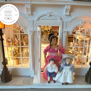 Puede incluir: Una tienda de mu&ntilde;ecas en miniatura con fachada blanca, puerta arqueada y dos farolas negras. Tres mu&ntilde;ecas est&aacute;n en la puerta. La tienda tiene un letrero que dice "Uptown Minis" y "Crafting Happiness".