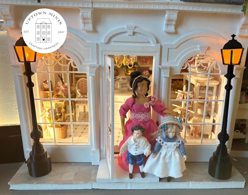 Puede incluir: Una tienda de mu&ntilde;ecas en miniatura con fachada blanca, puerta arqueada y dos farolas negras. Tres mu&ntilde;ecas est&aacute;n en la puerta. La tienda tiene un letrero que dice "Uptown Minis" y "Crafting Happiness".