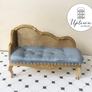 May include: A miniature gold chaise lounge with a blue fabric cushion and a woven cane back. The chaise lounge has ornate details and is sitting on a black and white tiled floor.