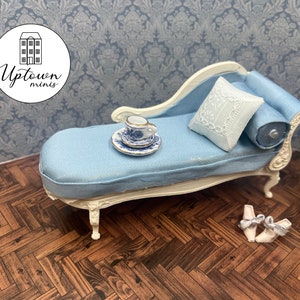 May include: A white chaise lounge with blue upholstery and a white pillow. A blue and white teacup and saucer are on the chaise lounge. The chaise lounge is on a wooden floor.