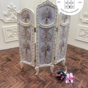 May include: A miniature three-panel folding screen with a floral design. The screen is cream-colored with ornate detailing and features a central panel with a vase of flowers. The screen is on a wooden surface. The Uptown Minis logo is in the upper right corner.