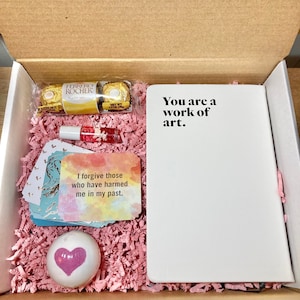 May include: A gift box filled with pink tissue paper and various items, including a small notebook with the text "You are a work of art." on the cover, a bath bomb shaped like a heart, a small bottle of nail polish, a card with the text "I forgive those who have harmed me in my past." and a box of chocolates.