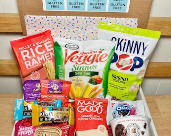 May include: A cardboard box filled with various gluten-free snacks. The box contains bags of rice ramen, veggie straws, popcorn, crackers, cookies, and a brownie. The box has a "Keep Calm It's Gluten Free" sticker.