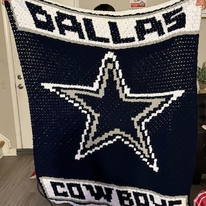 May include: A blue and white crocheted blanket with the Dallas Cowboys logo. The logo features a white star with a gray outline on a navy blue background. The words "Dallas Cowboys" are written in white with a gray outline below the star.