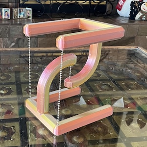 Puede incluir: Una escultura impresa en 3D con un diseño geométrico. La escultura presenta una base y una parte superior rectangulares, conectadas por piezas curvas y anguladas. El degradado de color transita del amarillo al naranja y al rosa. Cadenas plateadas suspenden los elementos superiores.