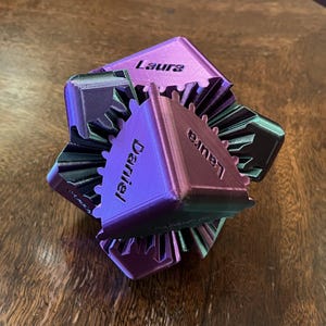 May include: A 3D-printed geometric puzzle in shades of purple and green, with the names "Laura" and "Daniel" printed on the faces. The puzzle has a gear-like design and sits on a wooden surface.