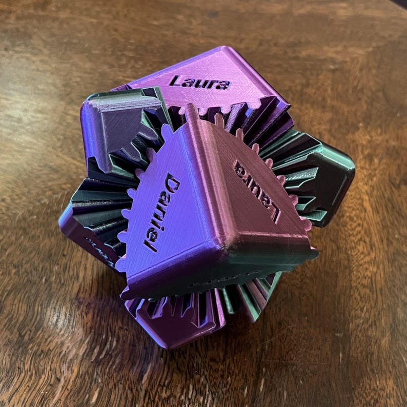 May include: A 3D-printed geometric puzzle in shades of purple and green, with the names "Laura" and "Daniel" printed on the faces. The puzzle has a gear-like design and sits on a wooden surface.
