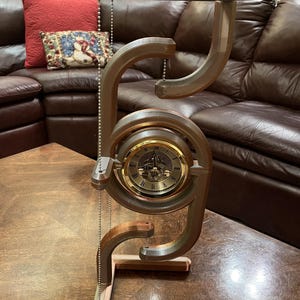 Puede incluir: Un soporte de reloj de metal de color cobre con una esfera redonda y una cadena que cuelga de la parte superior. La esfera del reloj tiene números romanos y un borde dorado. El soporte está sobre una mesa de madera.