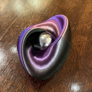 Puede incluir: Una escultura impresa en 3D con una forma arremolinada en forma de corazón. La escultura presenta un degradado de colores, pasando del morado al plateado y al negro. Una esfera plateada brillante está ubicada dentro de las curvas del diseño. El objeto está sobre una superficie de madera.