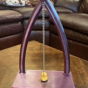 May include: A purple and gold perpetual pendulum with a silver chain and a gold weight. The base has the words "Perpetual Pendulum" in a yellow rectangle. The pendulum has an arch design.