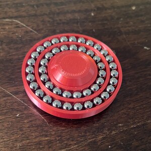 May include: A red 3D-printed fidget spinner with a circular design. The spinner has a central hub and two concentric rings filled with numerous small, shiny metal ball bearings. The design is simple and modern.