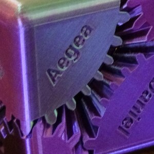 May include: Close-up of a purple, geometric puzzle with the name "Aegea" engraved on one of its sides. The word "Daniel" is also visible on another side. The puzzle has a gear-like design.