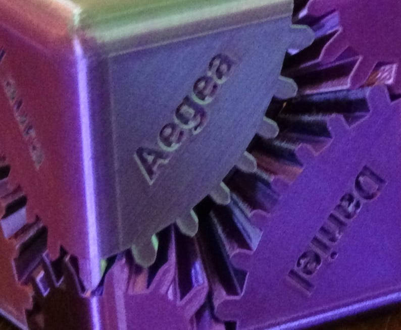 May include: Close-up of a purple, geometric puzzle with the name "Aegea" engraved on one of its sides. The word "Daniel" is also visible on another side. The puzzle has a gear-like design.