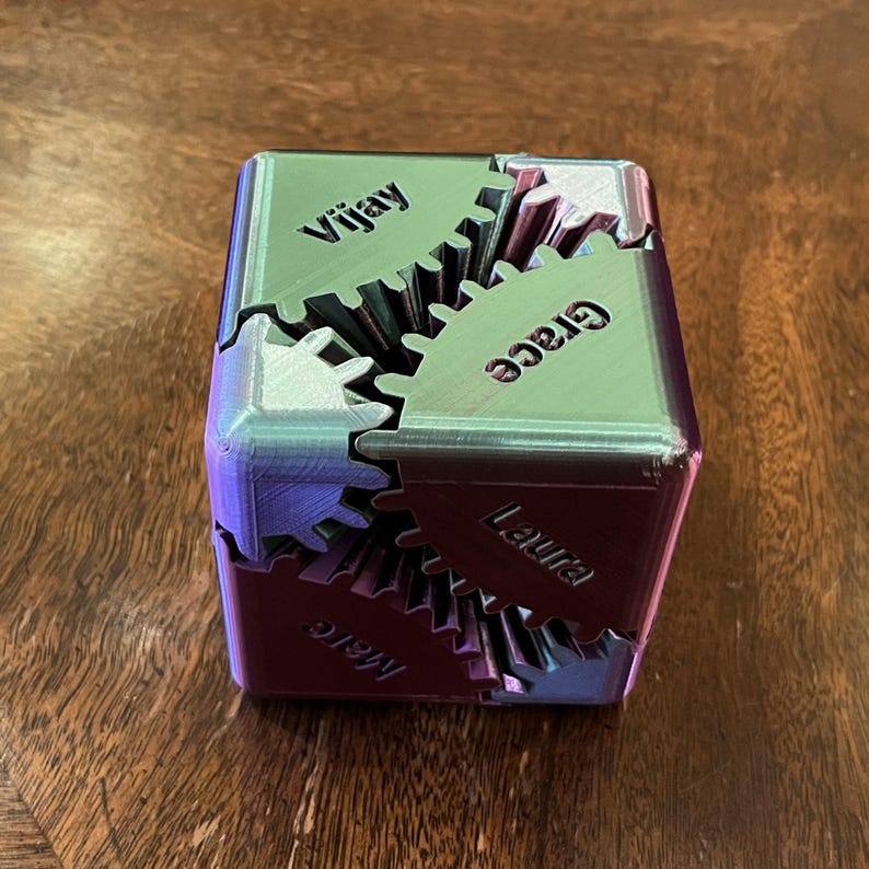 May include: A personalized cube puzzle with interlocking gear-shaped pieces. The cube is a mix of purple and green, with names "Vijay", "Grace", "Laura", and "Marc" printed on the sides. The puzzle is designed to be assembled.