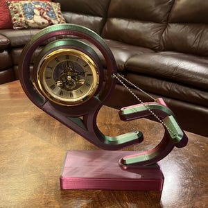 May include: A decorative clock with a gold-colored face and Roman numerals. The clock is set in a unique, swirling frame with a base, in shades of green, pink, and purple. A silver chain connects the frame.