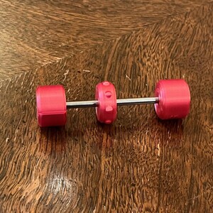 Puede incluir: Una pequeña pesa con una barra plateada y pesas de color rosa brillante. Las pesas son cilíndricas, con una pesa central texturizada. Esta pequeña pesa es probablemente un juguete o un artículo decorativo.