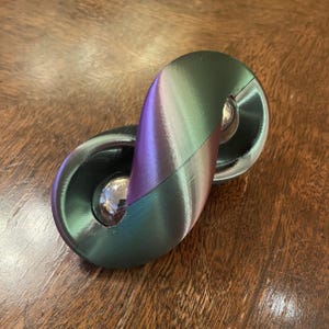 May include: A unique, iridescent fidget toy with a metallic sheen. The toy features two connected, rounded sections with a central, spherical bearing. The color shifts from green to purple, creating a visually appealing effect. The toy is designed for tactile engagement.