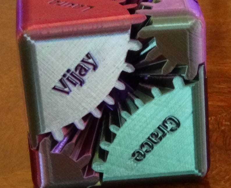May include: A multi-colored, 3D-printed gear cube with the names "Vijay" and "Grace" inscribed on adjacent gears. The cube features a mix of silver, red, and teal colors, showcasing the interlocking gear design.