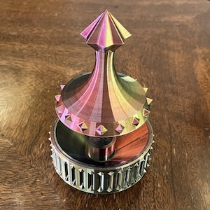 May include: A multi-coloured, metallic spinning top with a geometric design. The top features a conical shape with a pointed top and a circular base. The colours shift from pink to gold to green. The base has a decorative, ridged edge.