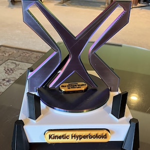 May include: A 3D printed kinetic sculpture of a hyperboloid, with a black and iridescent finish. The sculpture is mounted on a white base with a gold plaque that reads "Kinetic Hyperboloid". A smaller gold plaque on the sculpture reads "The Village Music by Sia Lee".