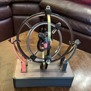 May include: A multi-colored kinetic sculpture with a complex, geometric design. The sculpture features a series of interconnected rings and supports, with a central, multi-faceted shape. The color palette includes pink, gold, black, and clear accents. The base is a dark gray.